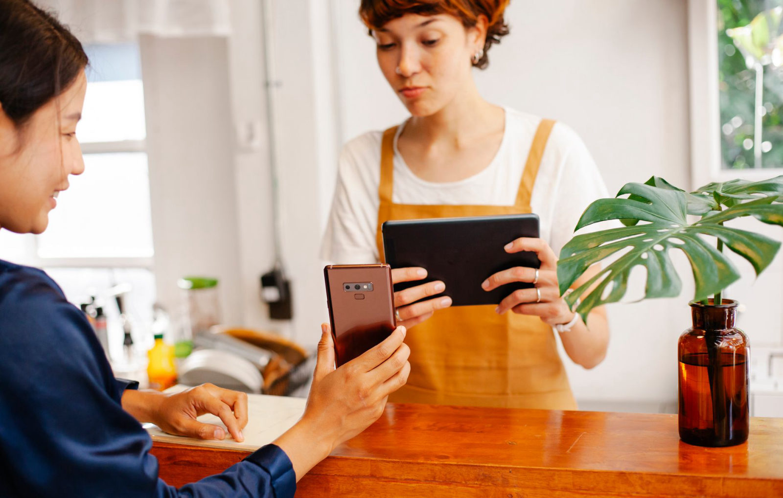 A café worker in a mustard apron reviews a tablet while a customer uses their smartphone at the counter — a relatable scene for small business owners wondering why is my small business not getting customers online